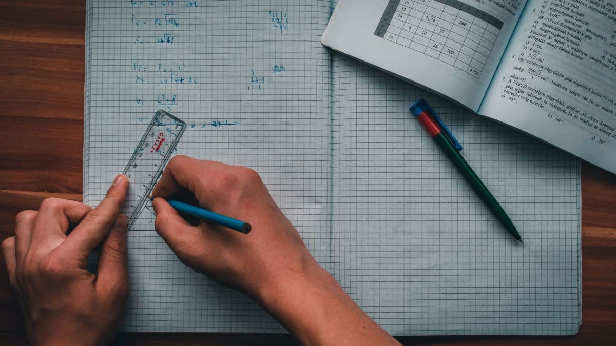 A person writes mathematical equations in a notebook using a ruler and pencil, focused on solving problems.
