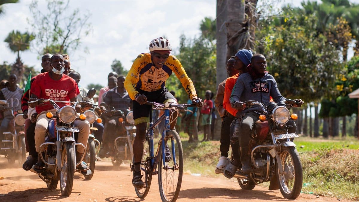 A dynamic race scene with a cyclist and boda boda riders on a dirt road, showcasing local culture.