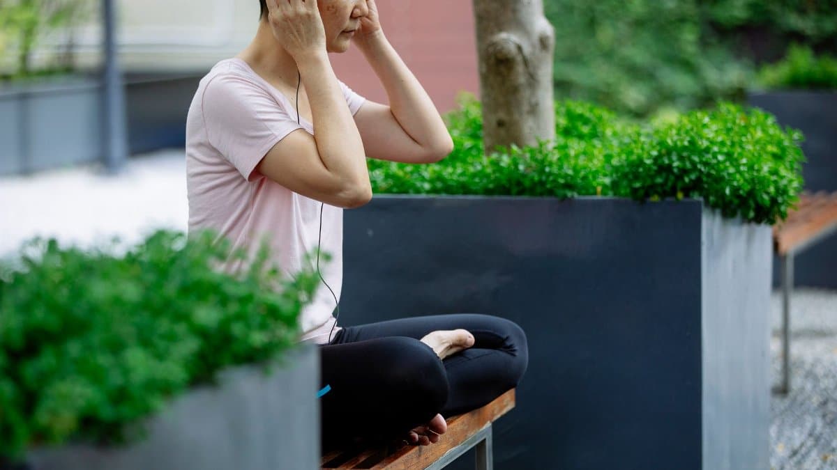 Crop middle age Asian woman in casual outfit and headphones sitting with crossed legs and closed eyes on bench in street near green plants and trees in pots in daylight