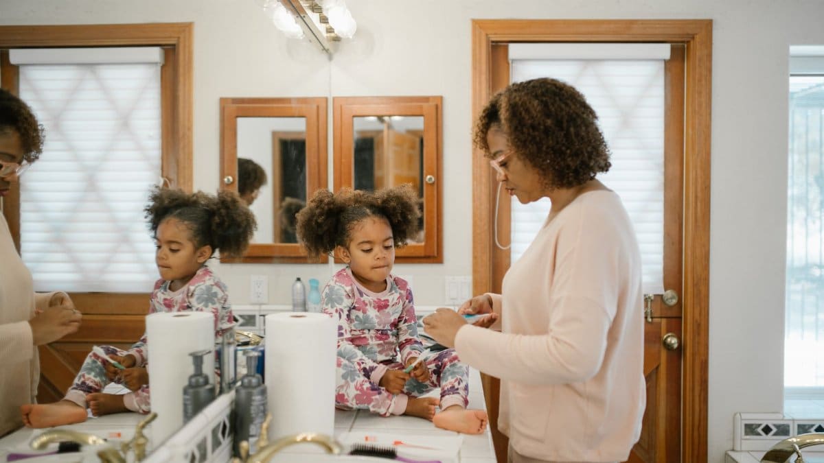 A mother assists her young daughter with her morning routine in a bright bathroom setting.