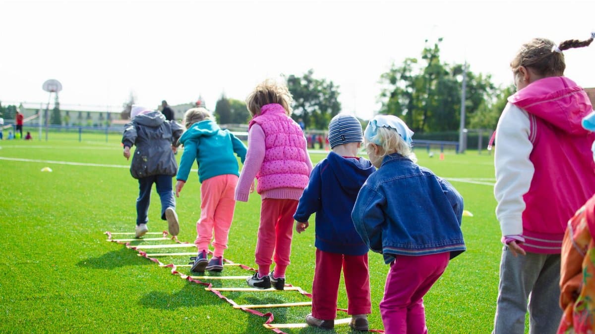 Children enjoy an outdoor activity on a grassy field, stepping over a ladder.