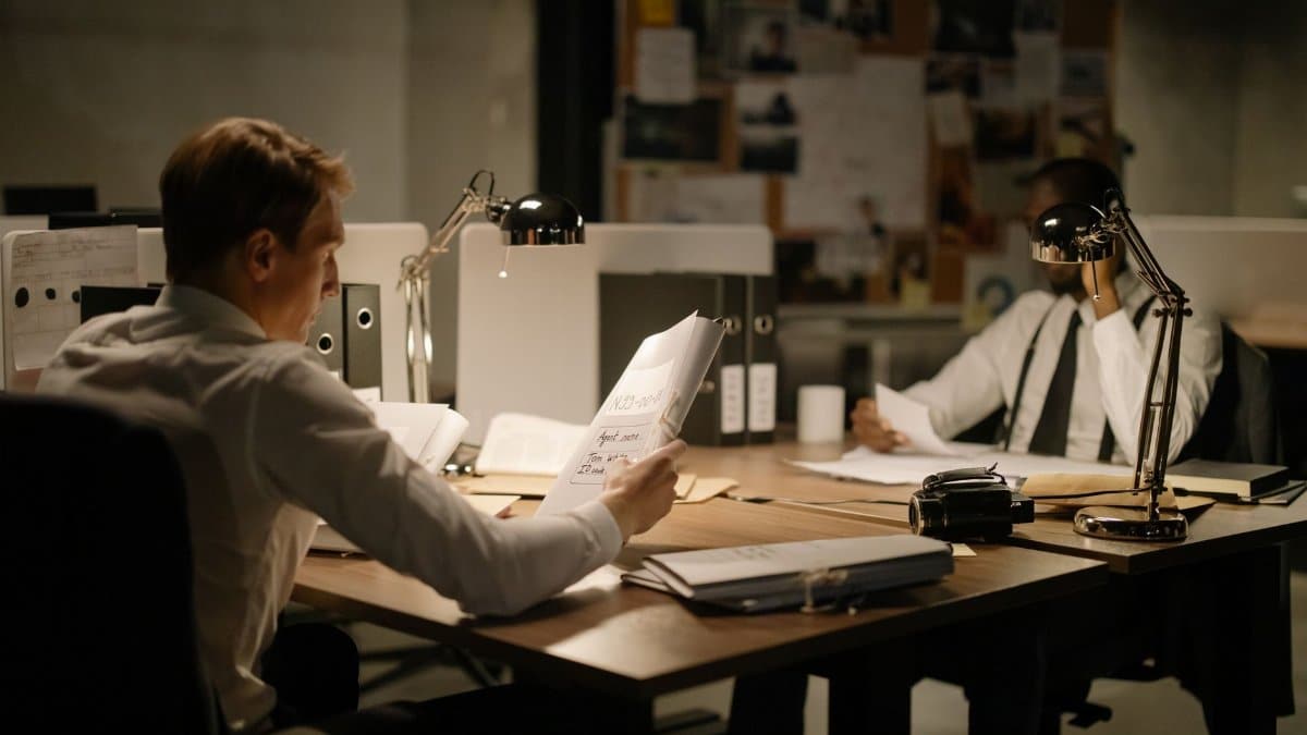 Two detectives analyzing documents in an office setting under dim lighting, focusing on investigation.