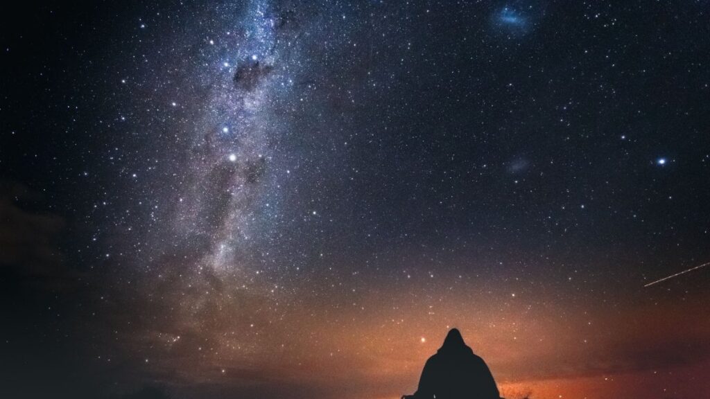 A lone figure sits under a starry sky with the Milky Way visible, capturing the beauty of a New Zealand night.