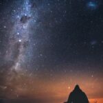 A lone figure sits under a starry sky with the Milky Way visible, capturing the beauty of a New Zealand night.