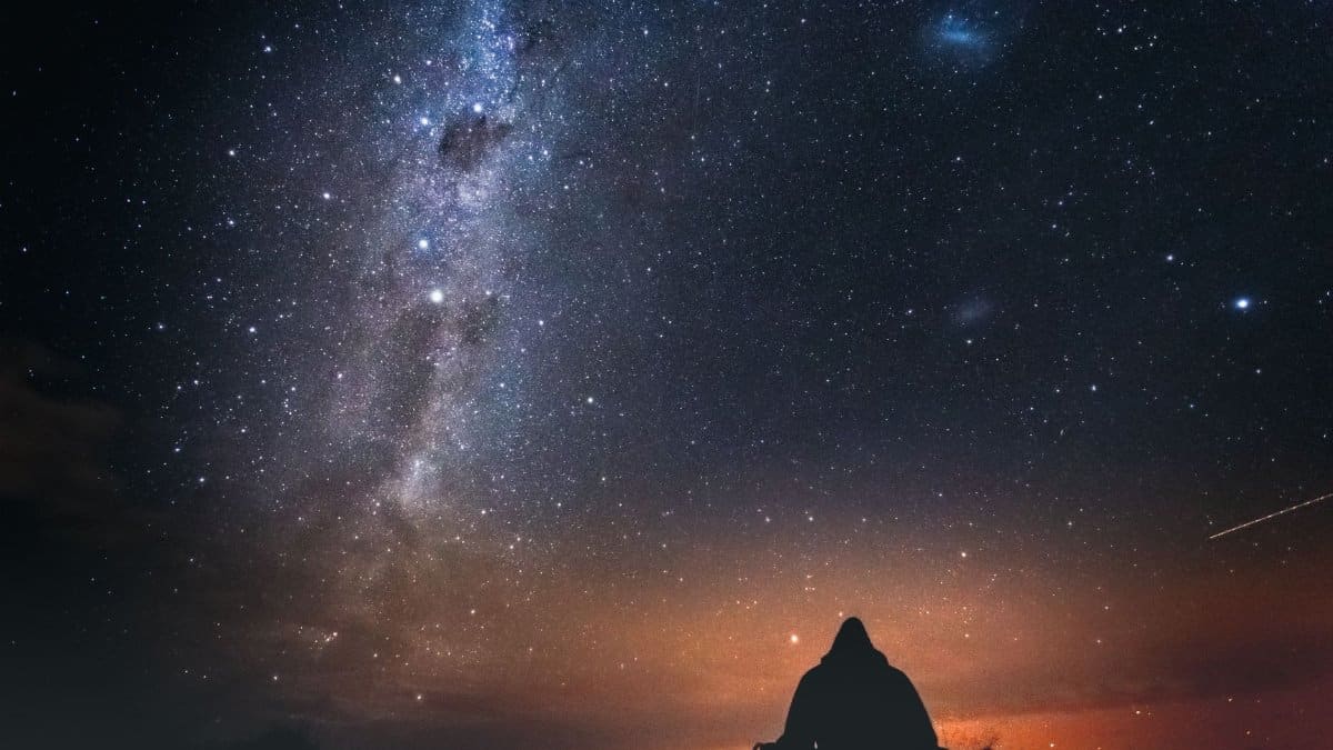 A lone figure sits under a starry sky with the Milky Way visible, capturing the beauty of a New Zealand night.