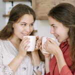 Two women smiling and enjoying hot beverages indoors, creating a warm, cozy atmosphere.