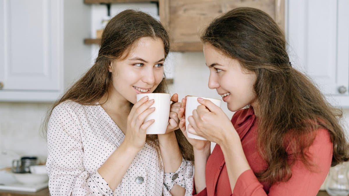 Two women smiling and enjoying hot beverages indoors, creating a warm, cozy atmosphere.