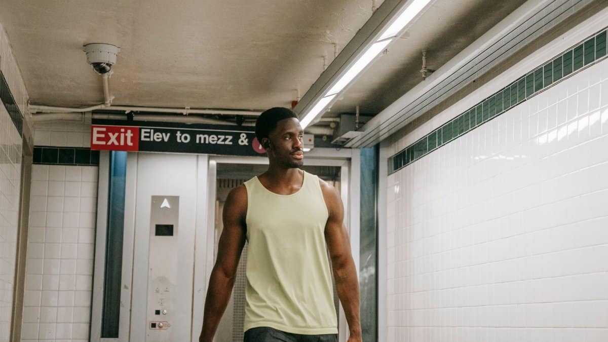 A man in a sleeveless top walks in a subway corridor near an exit sign, suggesting urban commute.