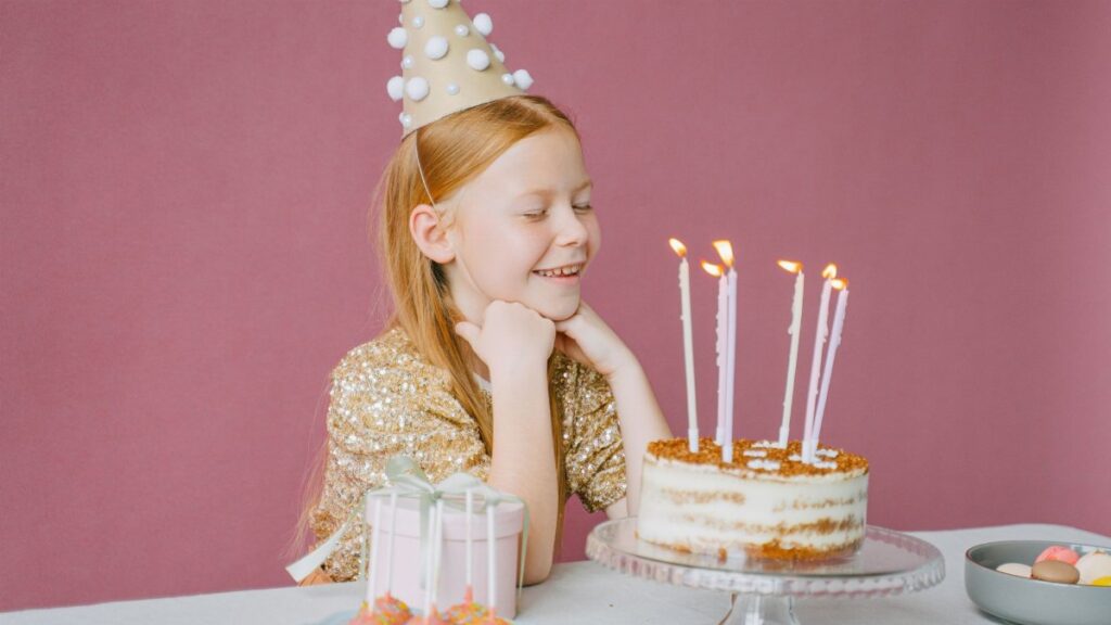 A young girl in a party hat smiles at her birthday cake with candles lit, ready to make a wish.