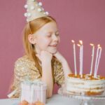 A young girl in a party hat smiles at her birthday cake with candles lit, ready to make a wish.