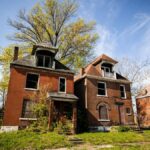 Three abandoned brick houses in a suburban neighborhood under a clear blue sky.