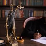 A focused judge writing on documents beside a Lady Justice statue in an office.