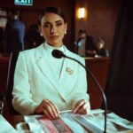 A poised woman in a white suit sits confidently in a courtroom setting, Baghdad.