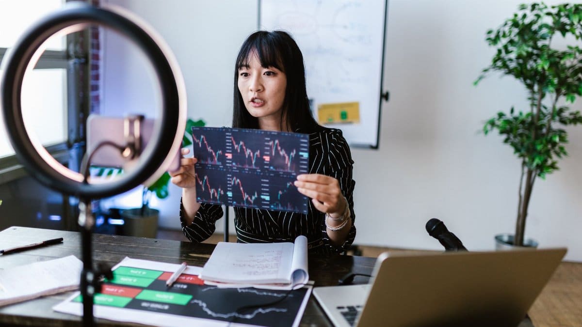 A woman vlogger discusses stock market trends using charts in a home studio.