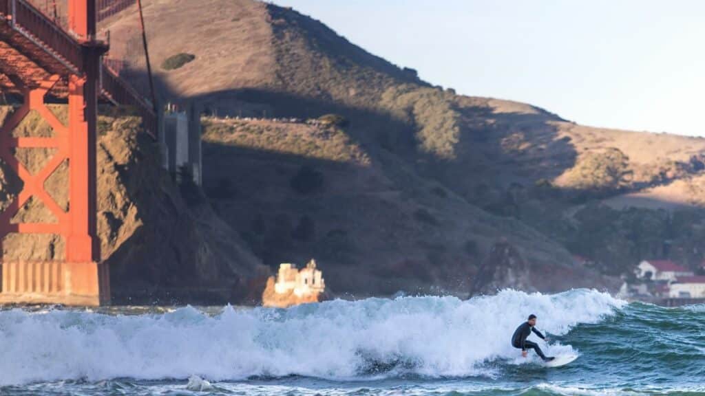 A surfer rides a wave near the iconic Golden Gate Bridge in San Francisco, California.