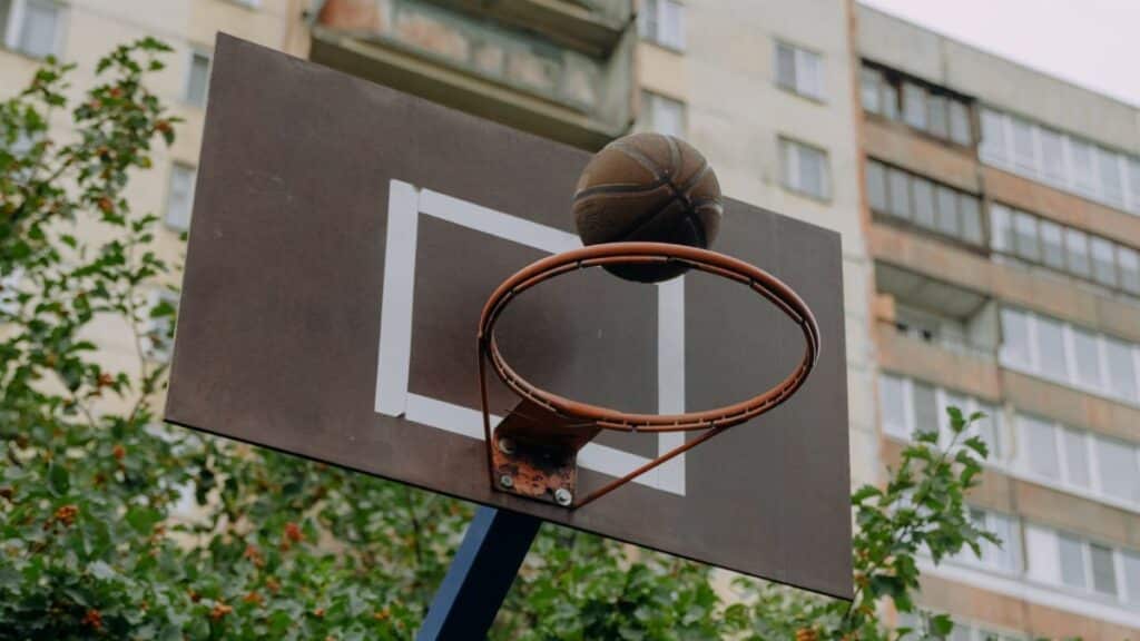 Close-up photo of a basketball hoop with backboard and ball in an urban setting.