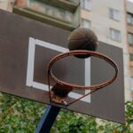 Close-up photo of a basketball hoop with backboard and ball in an urban setting.