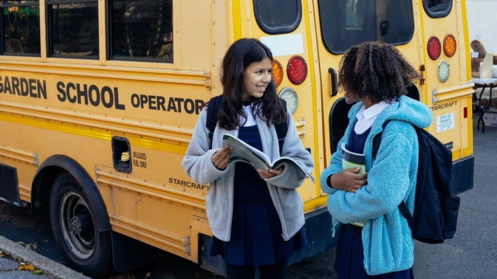 Happy multiracial classmates with backpacks standing near yellow school bus and communicating after school while looking at each other
