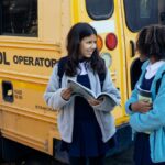 Happy multiracial classmates with backpacks standing near yellow school bus and communicating after school while looking at each other