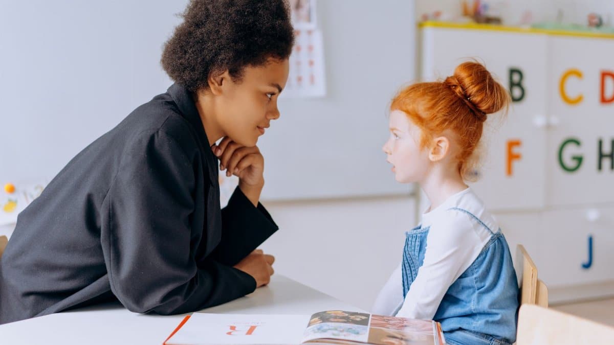 An adult teacher and a child student engage in a learning session at a classroom table.