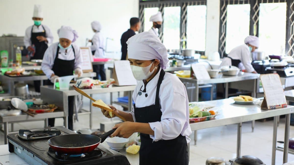 Chefs practicing culinary skills in a training kitchen setting, wearing masks for safety.