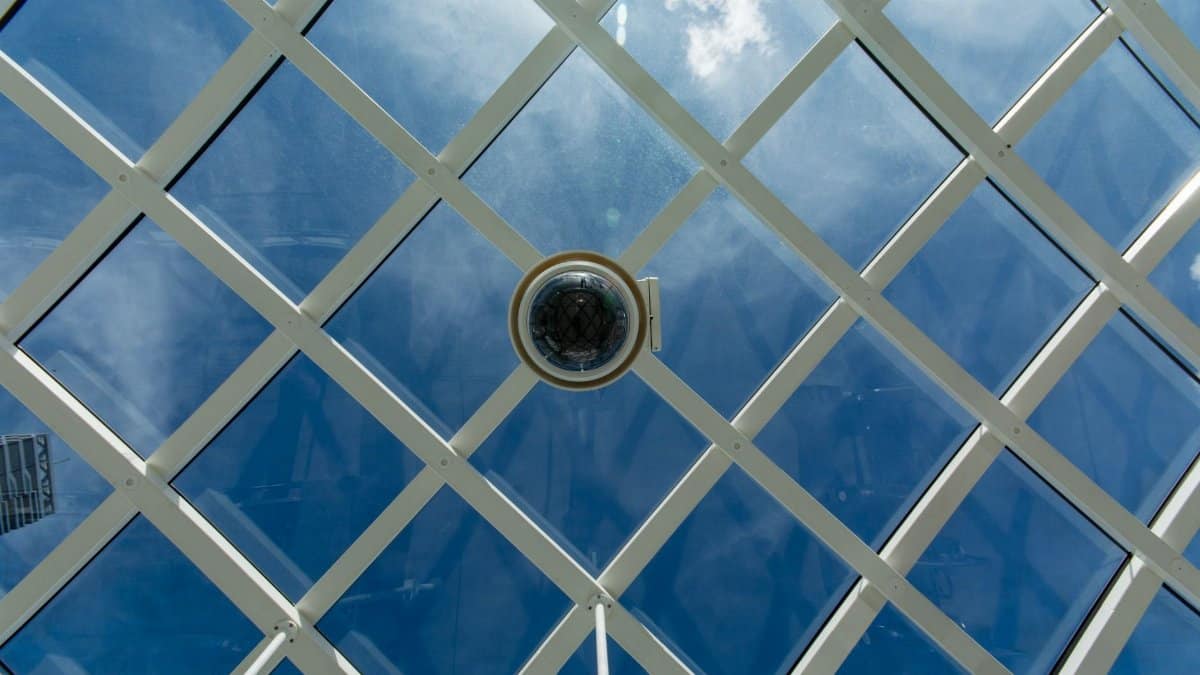 Surveillance camera mounted on a modern glass ceiling with sky reflections.