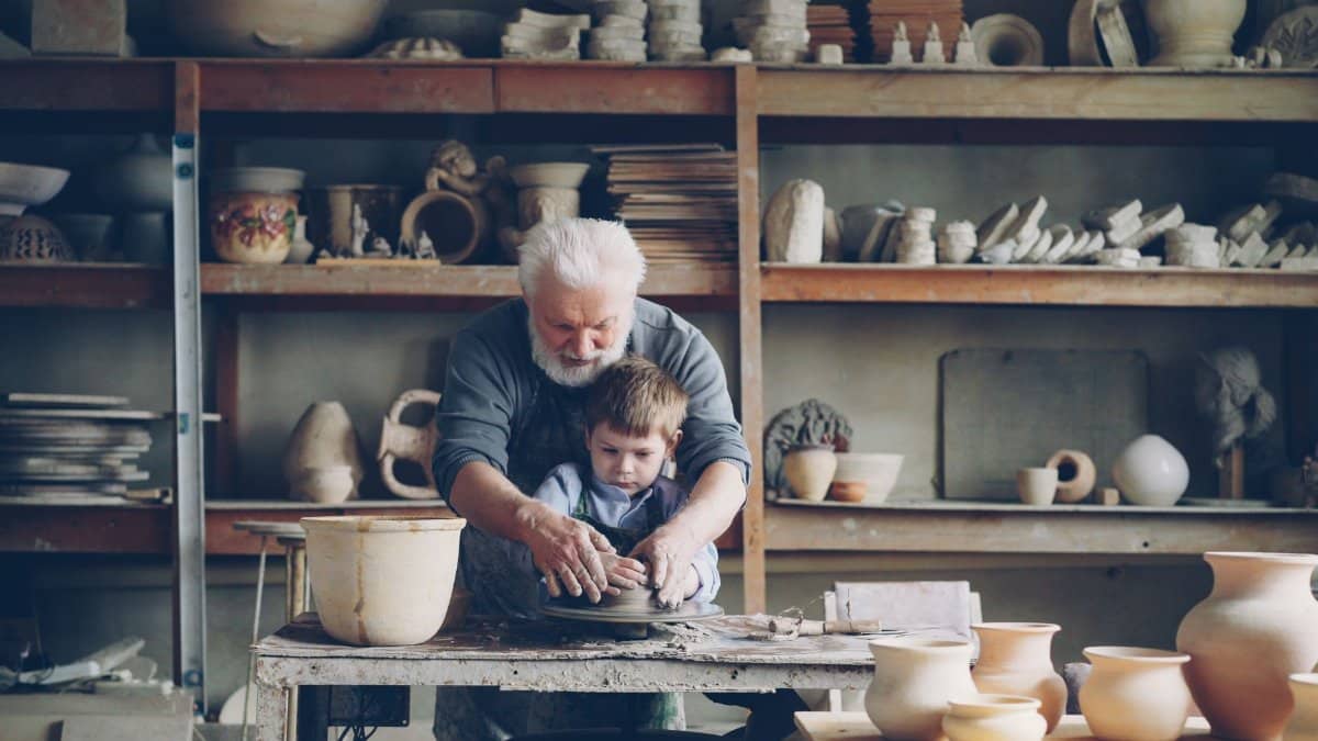 Elderly man teaching young boy pottery skills in a cozy workshop.