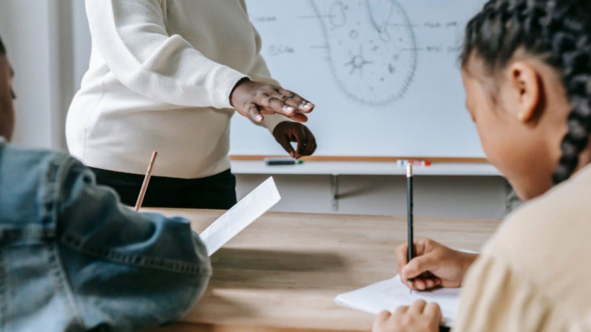 Crop faceless female teacher standing near desk while schoolchildren writing task on papers during lesson