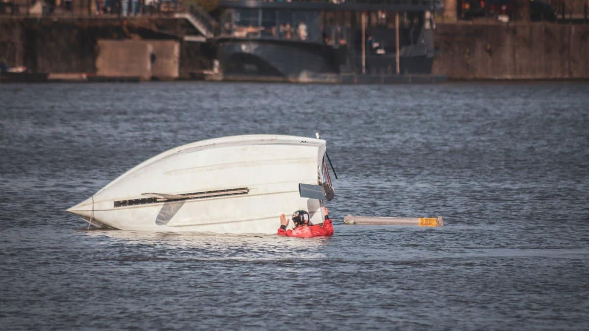 A capsized boat near an urban waterfront with a person wearing a life jacket visible in calm waters.
