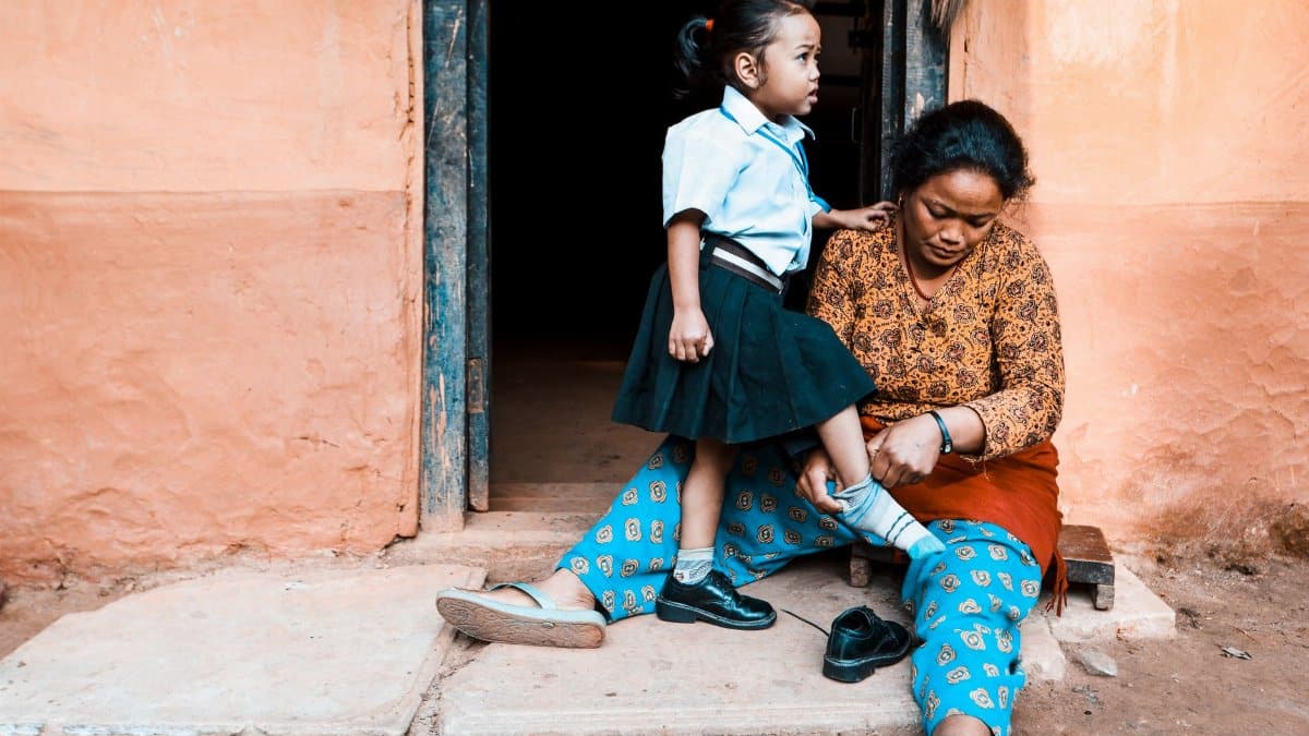 A mother helps her daughter put on socks before school, sitting outside their home.