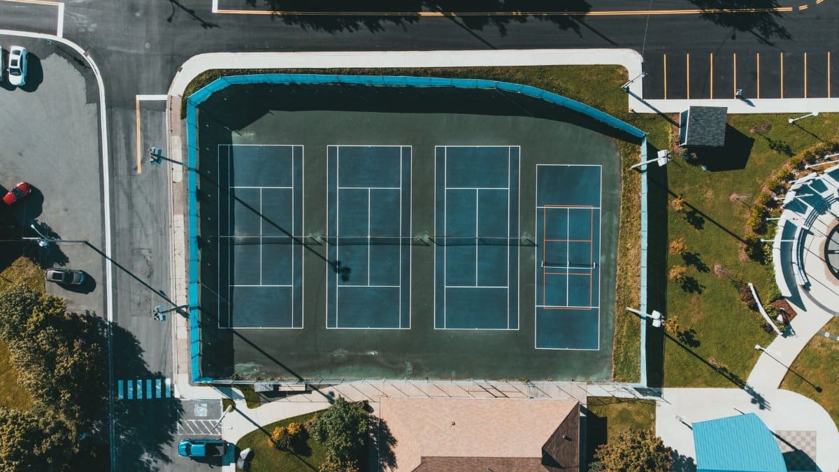 Aerial view showcasing tennis courts next to roads and parked cars in an urban setting.