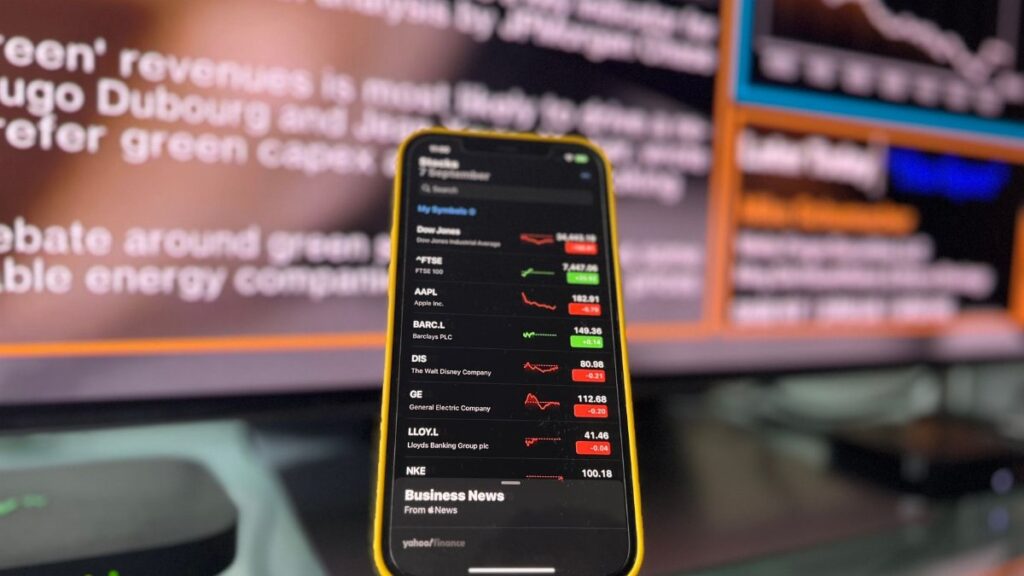 Smartphone showing stock market trends in front of blurred financial news screen indoors.