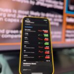 Smartphone showing stock market trends in front of blurred financial news screen indoors.