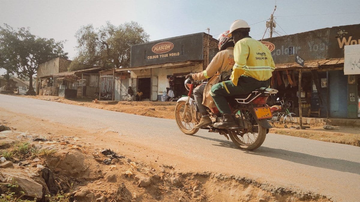 Two riders on a motorcycle traverse a dusty road in a small Ugandan town, lined with local shops.