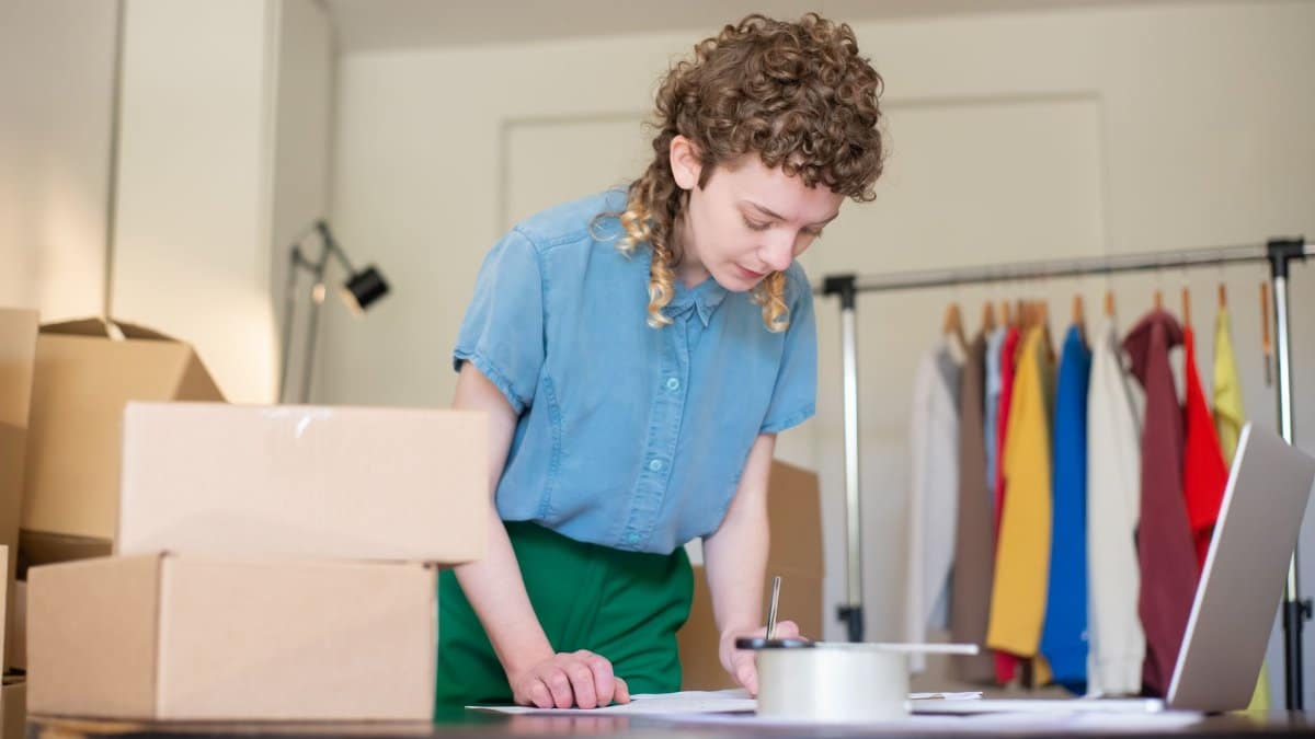 Woman packing online shop orders at home. Features boxes, clothing rack, and a laptop.