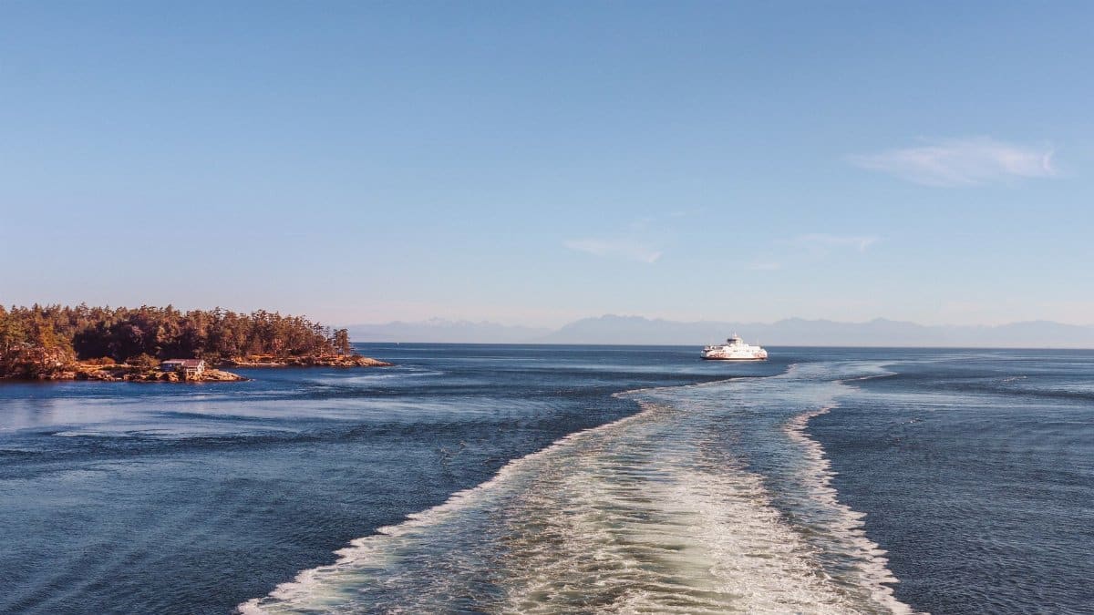 A ferry moves across calm sea near a wooded island under a clear blue sky.