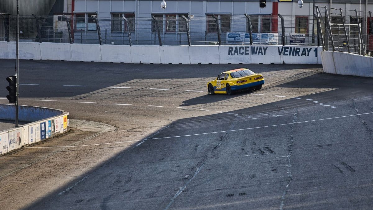 A yellow race car speeding on the track at Raceway Venray under clear skies.