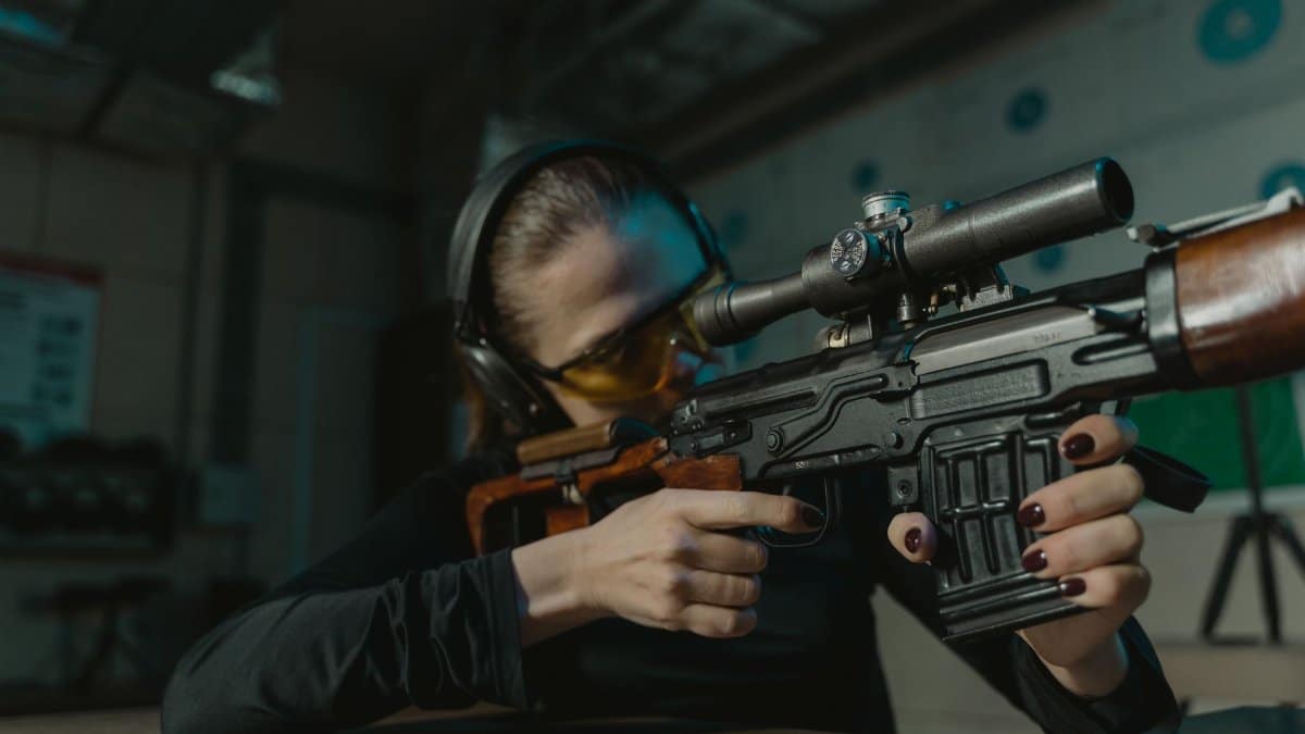 Woman in shooting range, wearing ear and eye protection, aiming a rifle with focus.