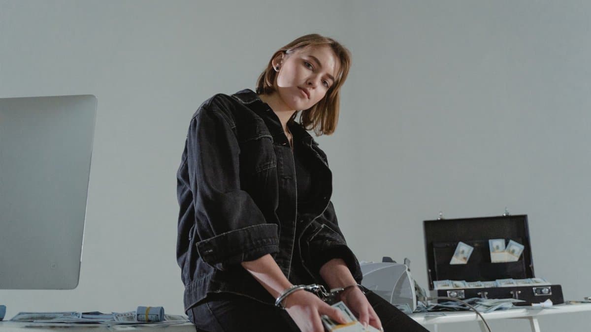 Young woman in black jacket handcuffed with cash, conceptual indoors setting.