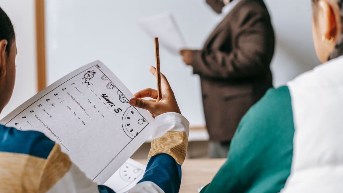 Multiethnic elementary classmates sitting at desk with copybooks and listening to black female teacher writing on whiteboard