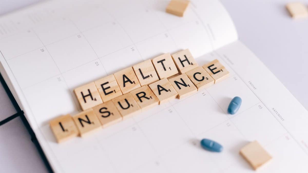 Close-up of health insurance spelled out with Scrabble tiles on a planner, alongside medication pills.