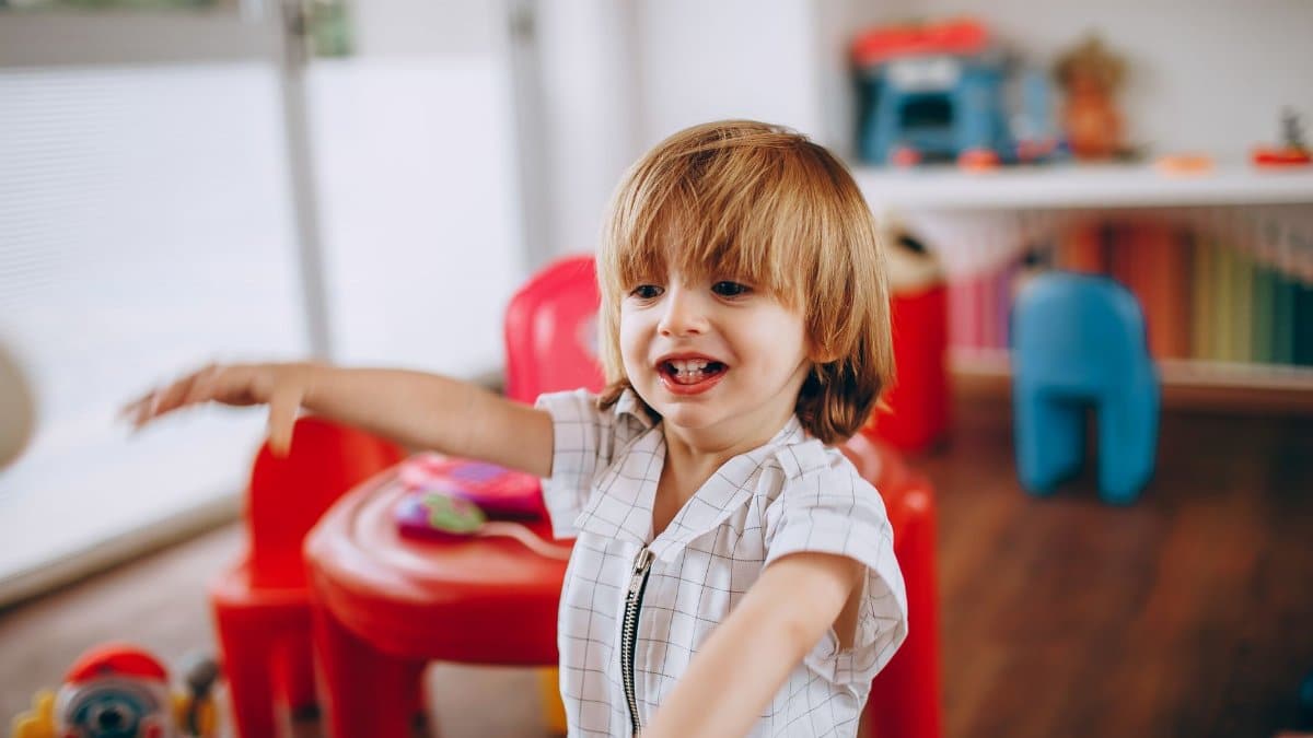 A cheerful child with long hair enjoys playtime in an indoor playground with red and blue plastic chairs.