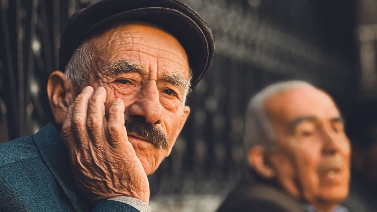 Elderly man in thoughtful pose wearing a cap, sitting outdoors in a sunny setting.