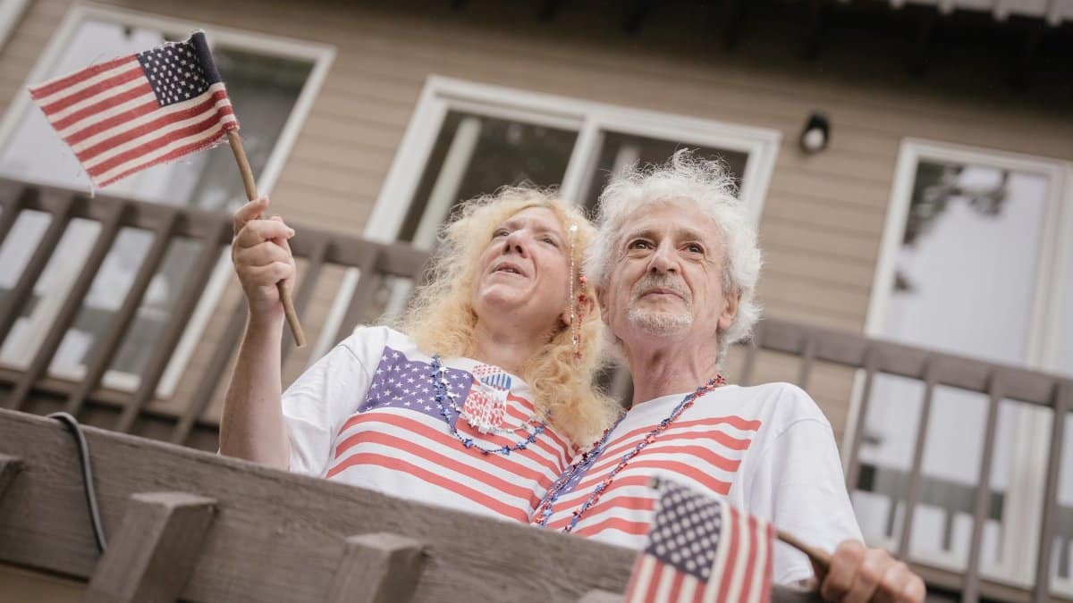 Elderly couple with American flags celebrating Fourth of July on a balcony.
