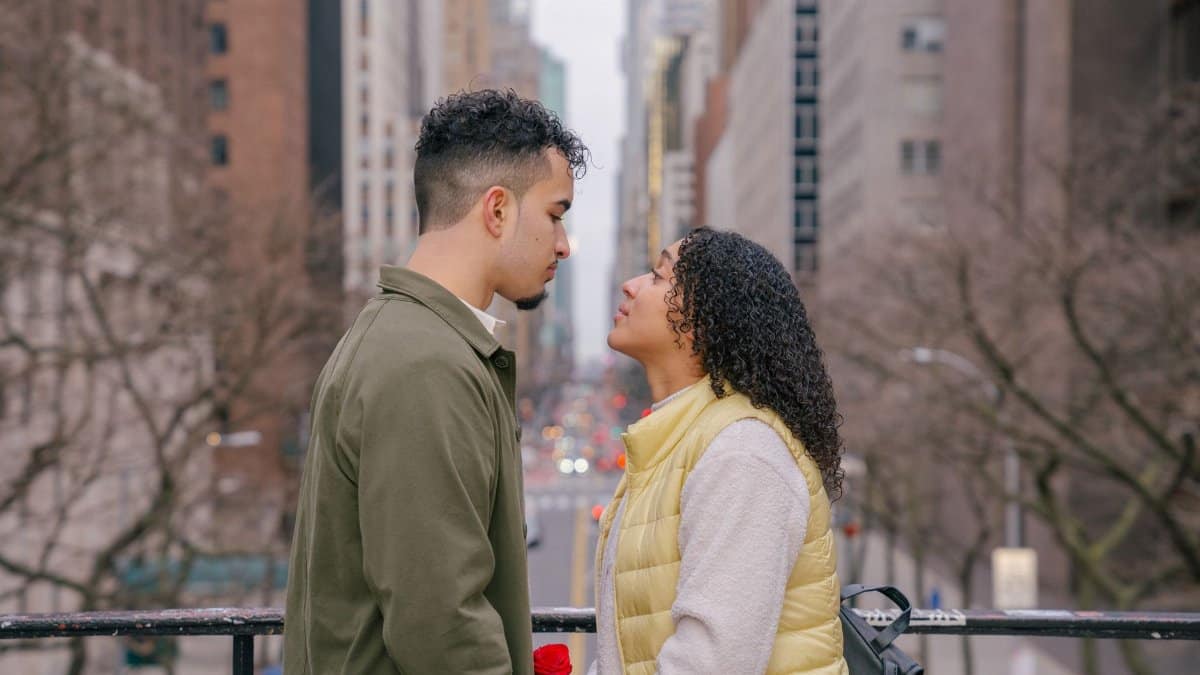 Side view of romantic Hispanic couple in casual clothes looking at each other while standing on bridge near metal fence against city street in daytime