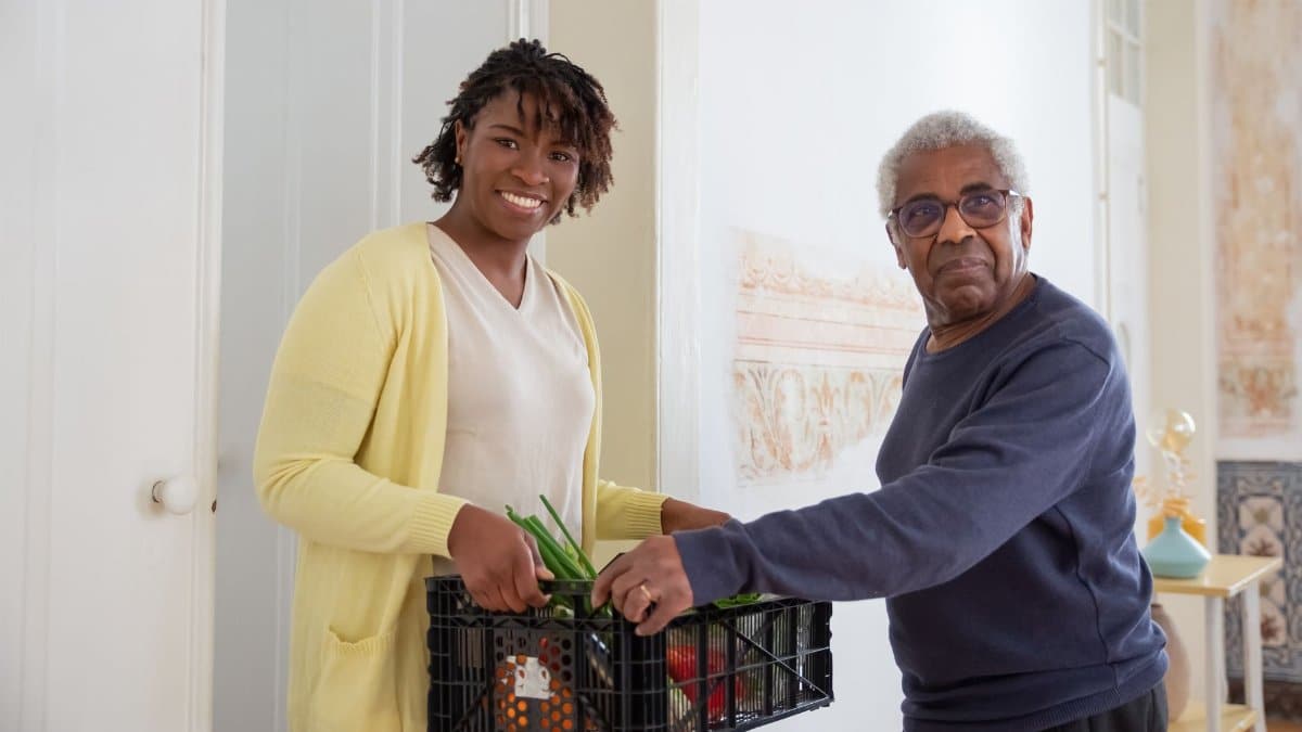 Smiling woman assists a senior man with food delivery indoors, promoting community support and care.