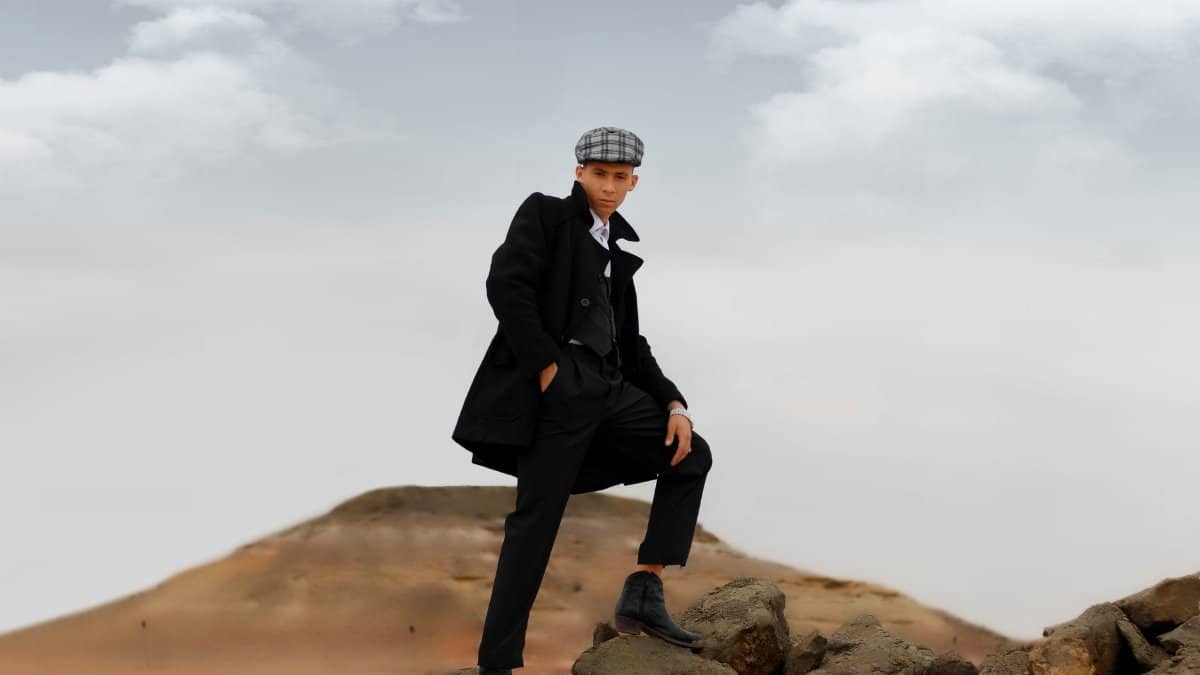 Young man in a stylish coat and cap poses on rocky terrain under cloudy sky. Fashionable and confident.