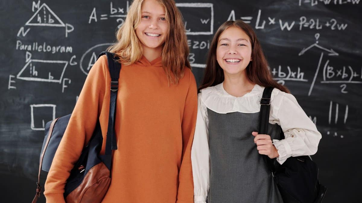 Two smiling students with backpacks standing in front of a chalkboard filled with math formulas.