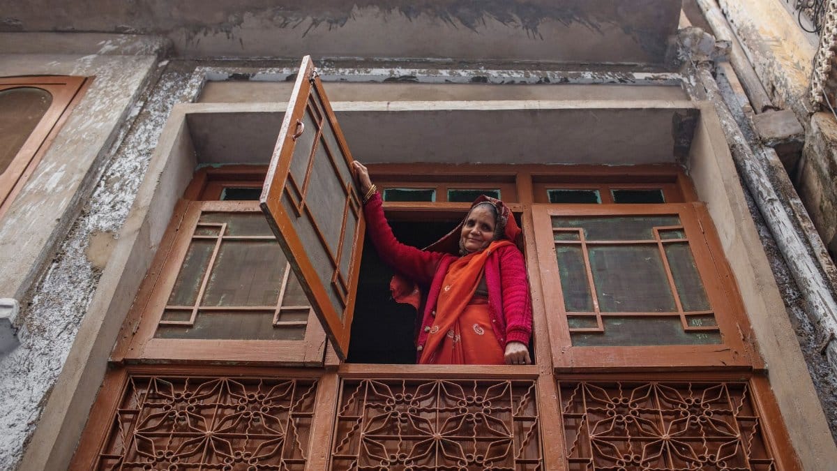 From below of cheerful senior ethnic female in traditional colorful sari looking at camera opening window of oriental balcony