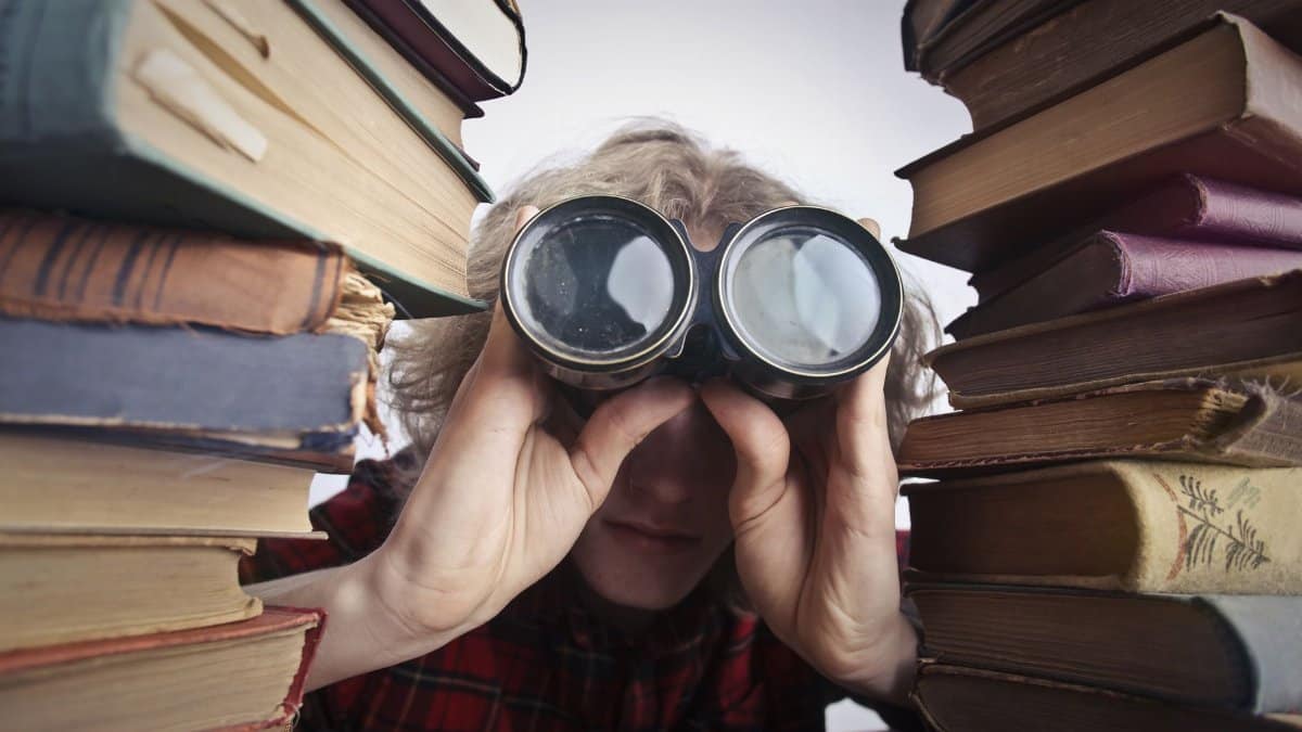 Young unrecognizable person sitting at table with stacked old books and looking through binoculars